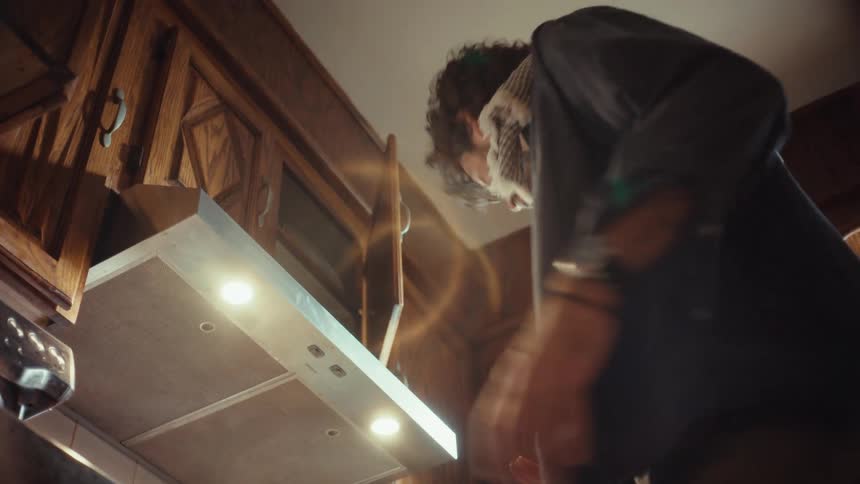 A dramatic low-angle shot captures a man intently cooking, stirring a pan on a modern gas stove