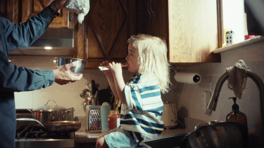 A father and son playfully cook together in slow motion in their messy kitchen, laughing as they toss ingredients into a bowl