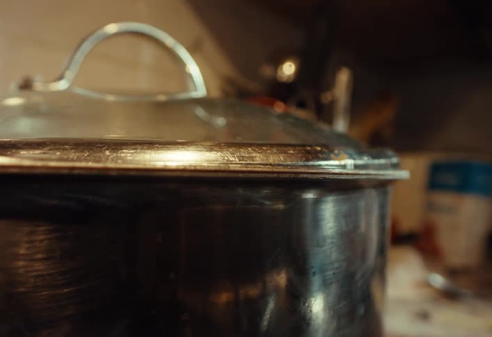 A hand places a glass lid on a steaming stainless steel pot while cooking in a kitchen