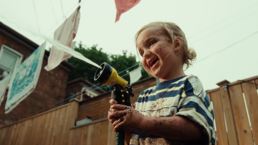 A gleeful muddy toddler laughs while spraying a garden hose in the backyard in super slow motion