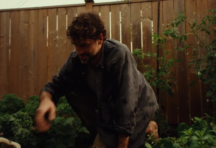 A playful, muddy boy throws dirt at his father before the man begins watering the lush vegetable garden