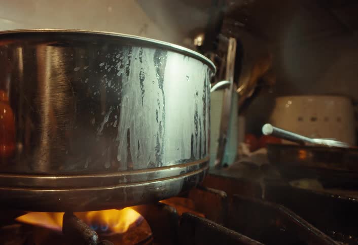 A close-up shot of a stainless steel pot steaming and boiling on a gas stove in a busy restaurant kitchen