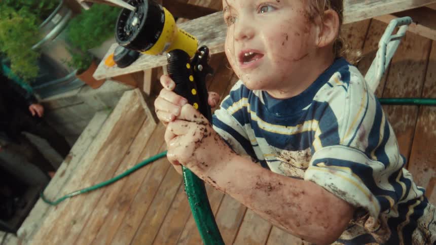 A close-up slow motion shot shows a muddy toddler with a look of pure excitement spraying a garden hose nozzle