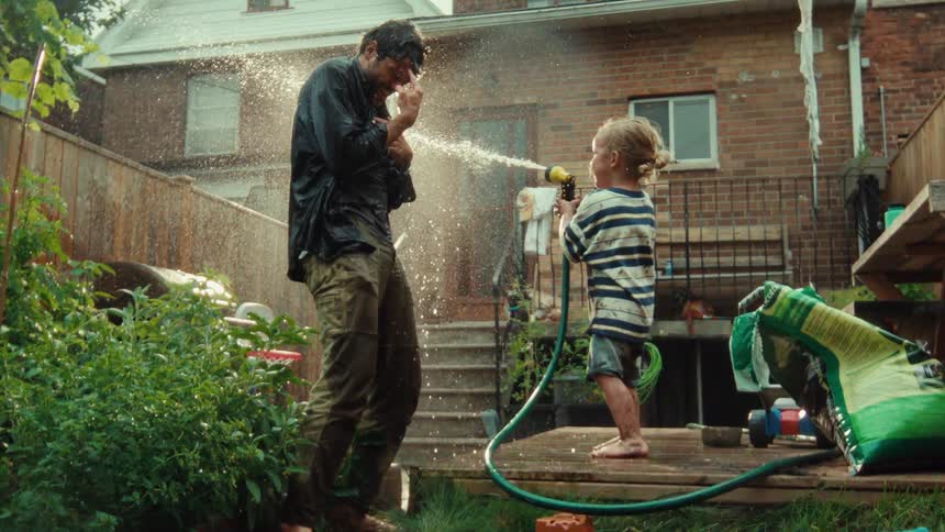 A laughing toddler playfully sprays their father in the face with a garden hose in the backyard, captured in slow motion