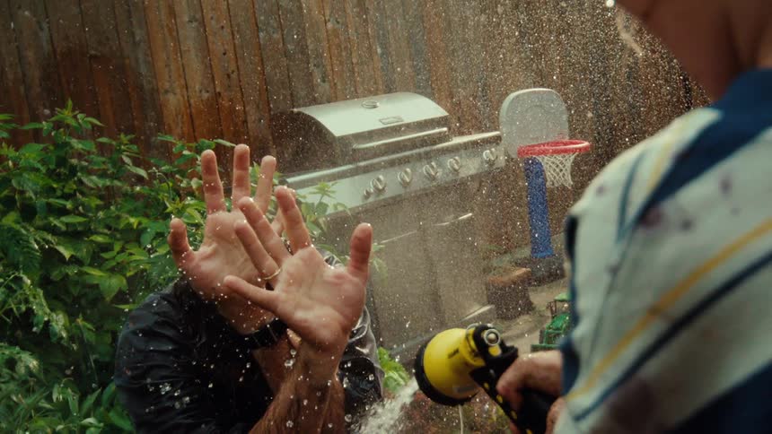 A young boy playfully sprays his father in the face with a garden hose in their backyard, captured in super slow motion
