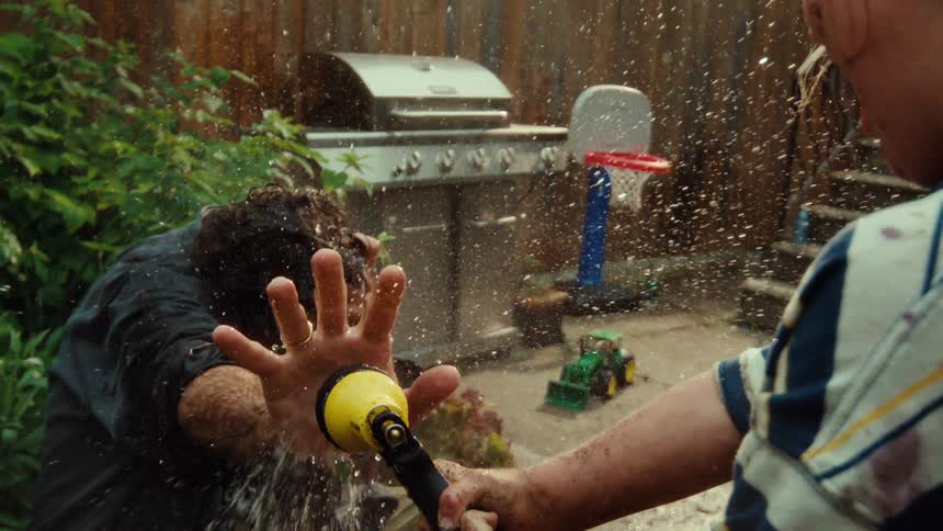 A father gets sprayed in the face with a garden hose by his son in the backyard during a playful moment in slow motion