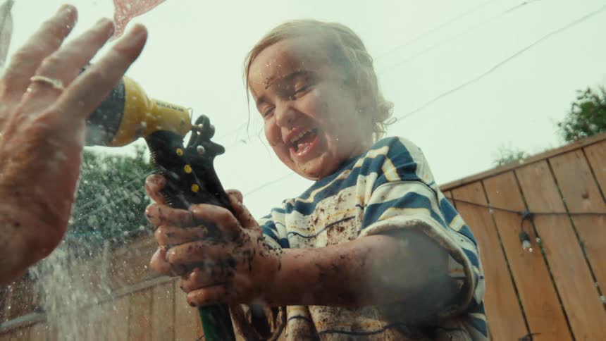 A happy toddler covered in mud laughs as he sprays a garden hose at his parent in the backyard, captured in slow motion