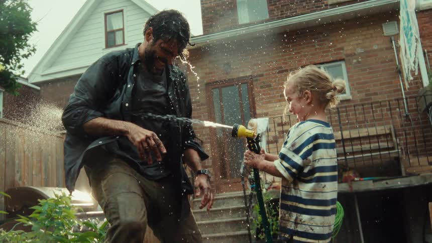 A joyful young girl playfully sprays her muddy father with a garden hose in their backyard in cinematic slow motion