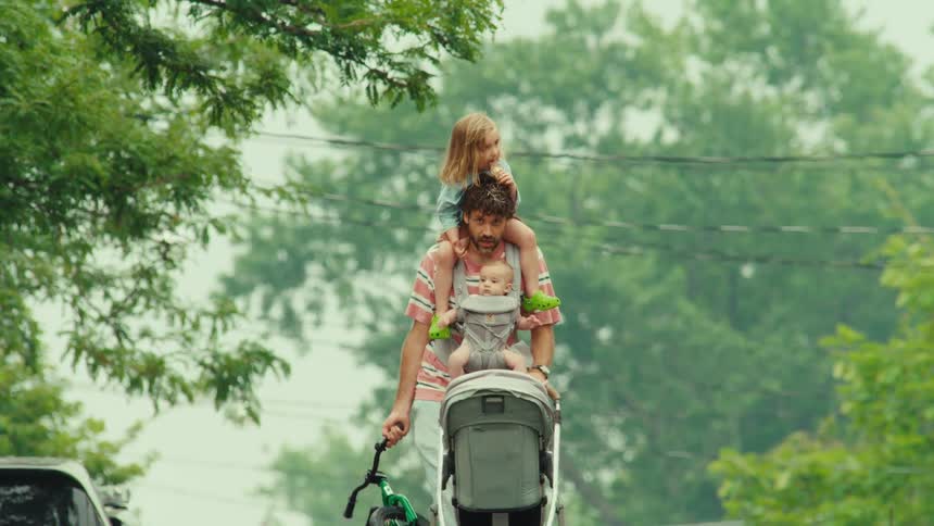 A multitasking father walks down a leafy suburban street with his toddler on his shoulders, a baby in a carrier, pushing a stroller and carrying a small bike