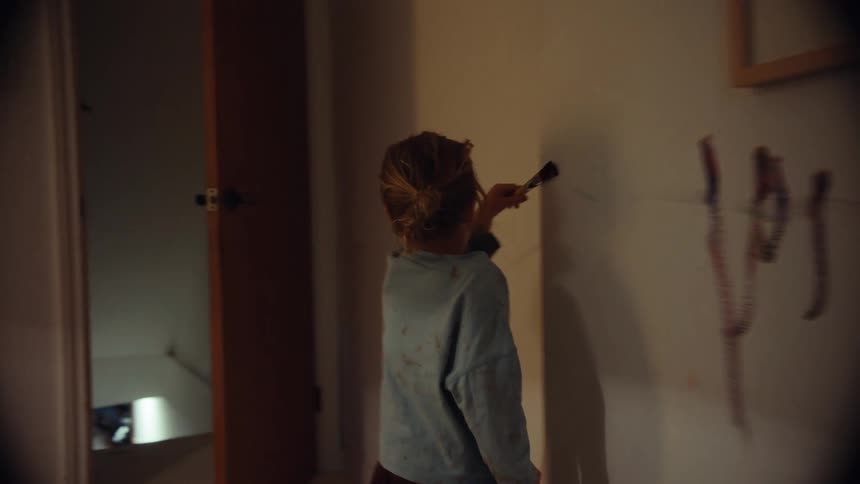 A mischievous young girl with paint on her face happily paints on the wall of her home