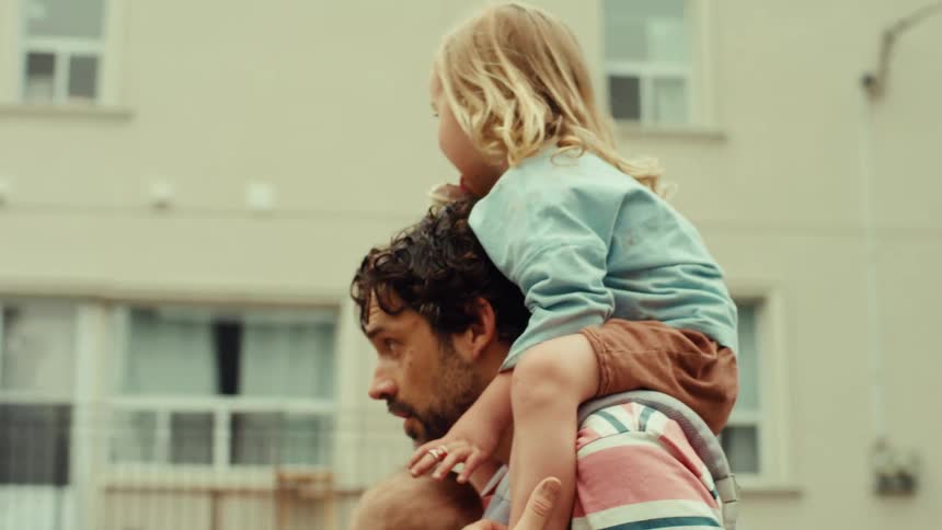 A loving father multitasks as he carries his baby in a front carrier and gives his toddler a shoulder ride while walking through the neighborhood