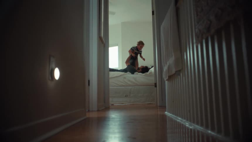 A father playfully lifts his young child in the air while lying on a bed in a sunlit bedroom, viewed from a hallway