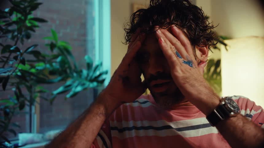 An exhausted artist covered in paint rubs his face in frustration while sitting in his studio