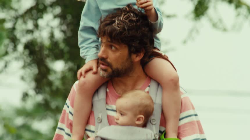 A father takes a walk carrying his baby in a carrier and toddler on his shoulders while she eats a messy ice cream cone