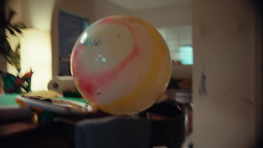A stressed-out man with paint on his face sits at a messy craft table, looking overwhelmed as a beach ball floats by