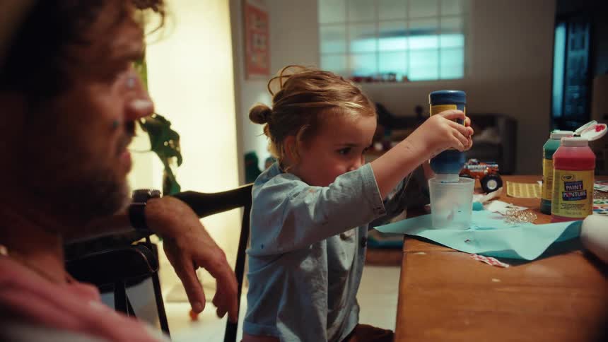 A sweet young girl intently helps her father squeeze blue paint into a cup during a fun arts and crafts session at their dining table