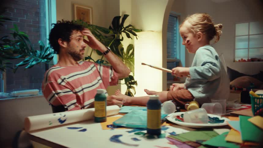 A joyful father and his young daughter laugh and playfully paint each other while doing arts and crafts at their messy dining table