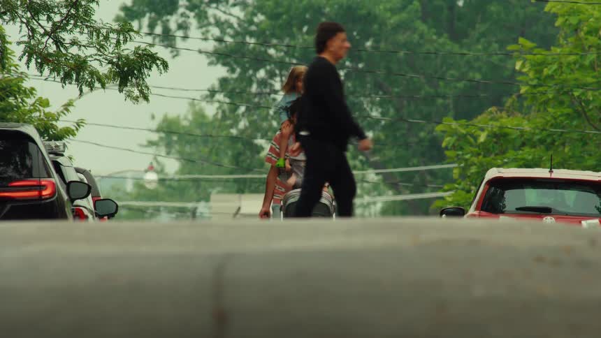A father walks down a suburban street with his toddler on his shoulders and a baby in a carrier while pulling a stroller