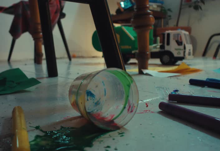 A chaotic scene unfolds as a cup of paint spills and markers fall onto the floor during a messy arts and crafts session