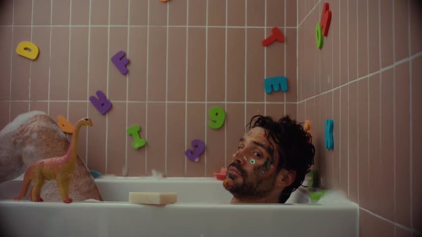 A despondent man with paint on his face sits in a bubble bath filled with children's toys before sinking beneath the water
