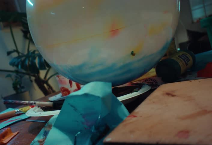 Close up of an abstract painted sphere rolling across a messy artist's desk with art supplies