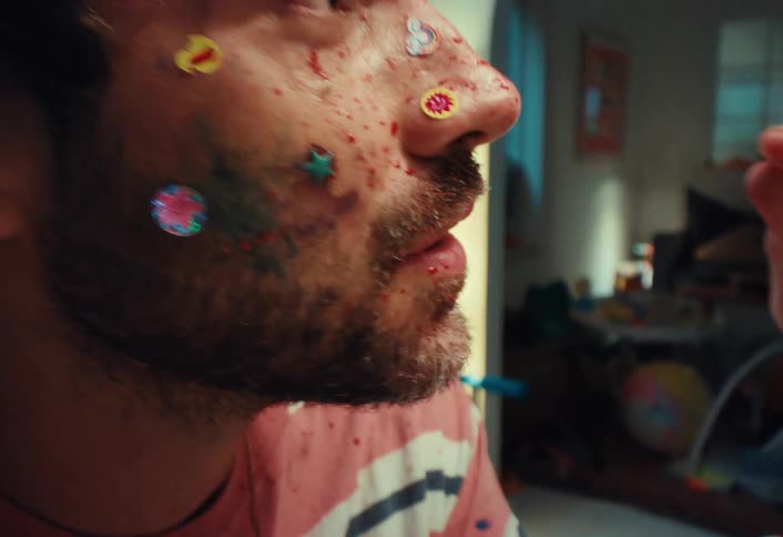 An exhausted father rubs his eyes, his face a canvas of children's stickers and messy paint