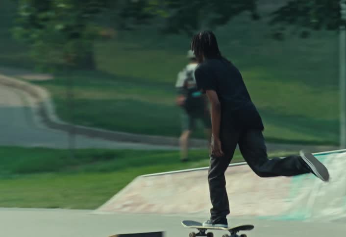 A young man with long hair smoothly glides across a graffiti-covered skatepark in slow motion