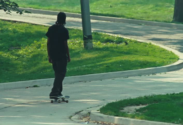 A person seen from behind rides a skateboard in slow motion down a paved path in a park, then picks up the board and walks away