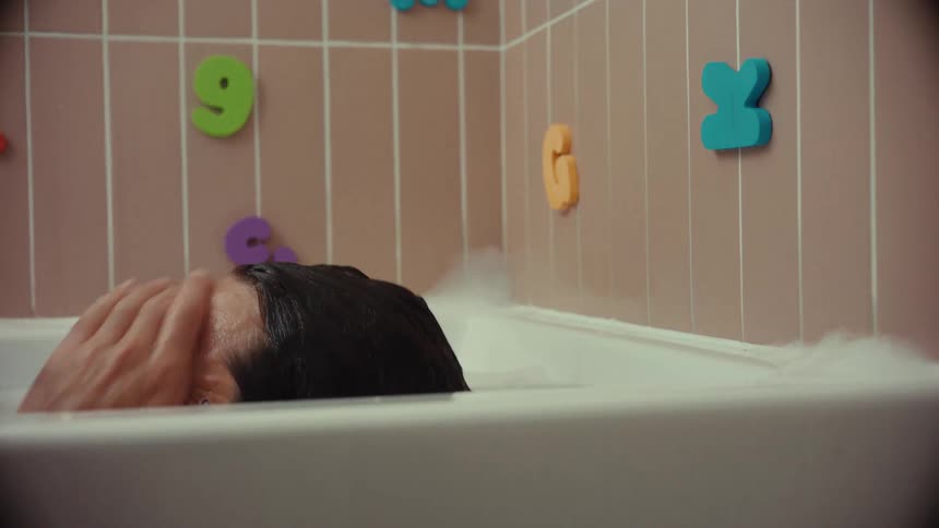 A stressed and exhausted father with stickers on his face slowly submerges himself in a bubble bath to decompress