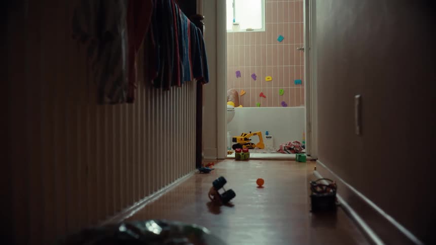 A stressed father tries to relax in a bathtub at the end of a hallway cluttered with children's toys
