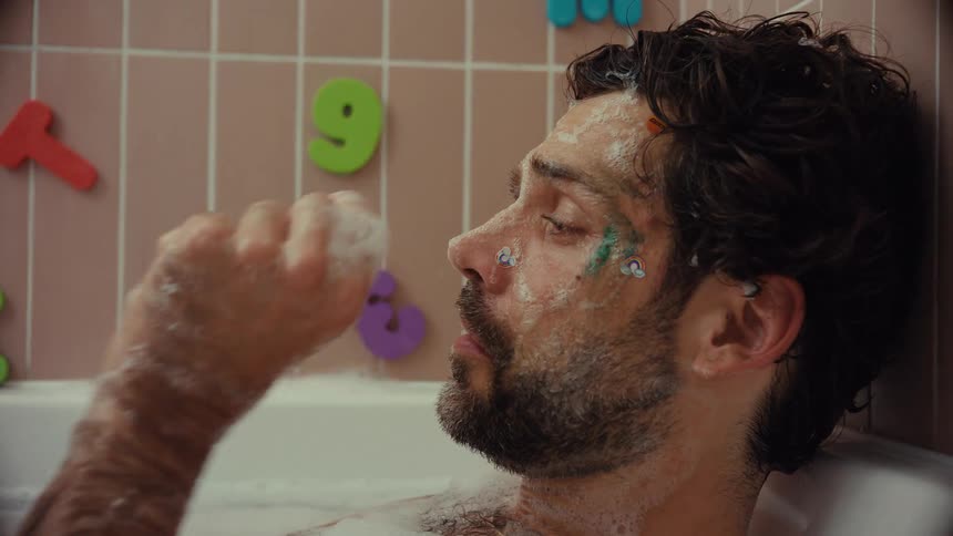 Close up of an exhausted father covered in soap, glitter, and stickers relaxing in the bathtub after playing with his kids