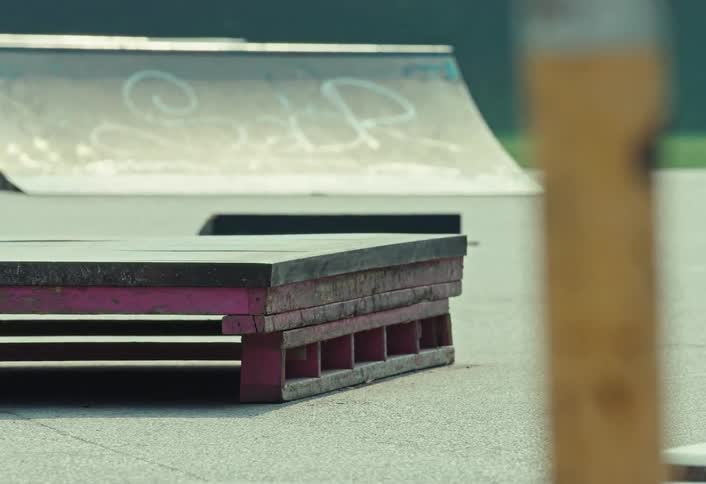 A static shot shows a pink grind box and a ramp with graffiti at an empty outdoor skatepark on a sunny day