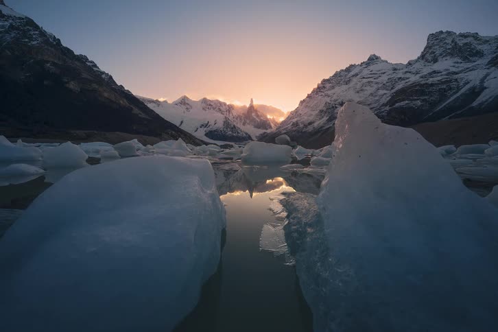 Serene time-lapse pulls back from glowing icebergs to reveal a majestic sunset over a glacial lake surrounded by snowy mountains