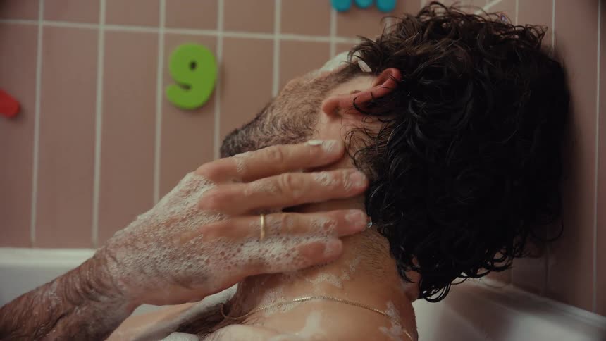 A tired man with stickers and paint on his face slowly washes himself in a soapy bubble bath