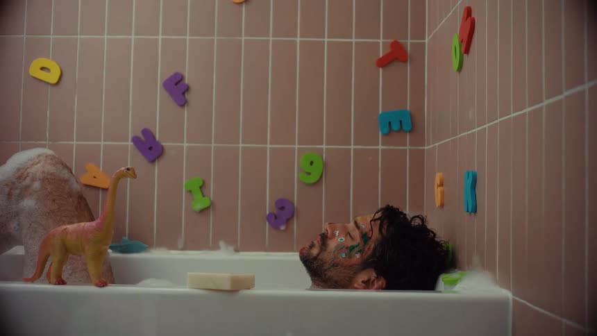 An exhausted father with a painted face falls asleep in a bubble bath surrounded by children's toys, slowly sinking before waking up abruptly