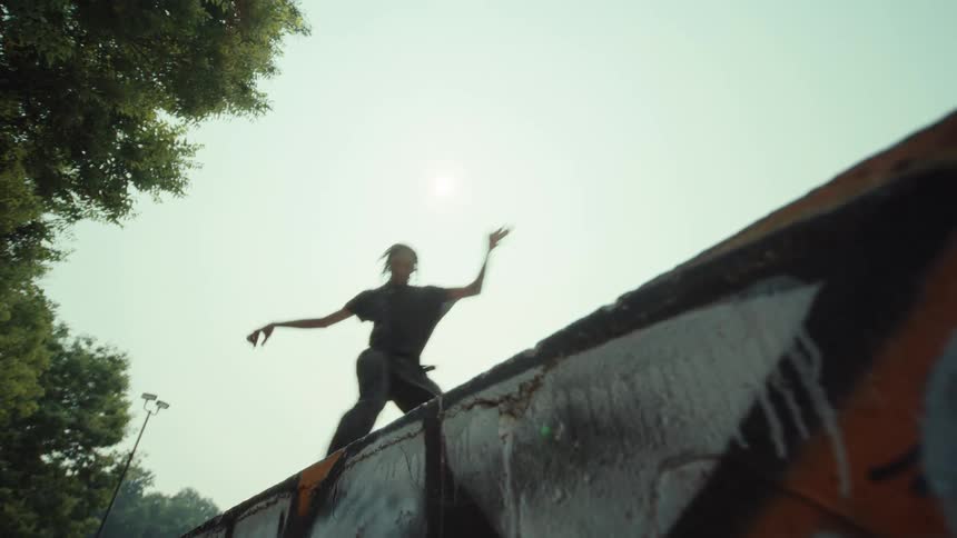 A person with dreadlocks runs and dances expressively along the top of a graffiti-covered wall against a bright sun