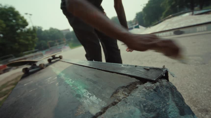 A close-up slow-motion shot of a skateboarder's hands waxing a metal ledge at an outdoor skatepark