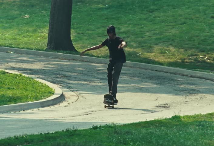 A young man wearing all black cruises down a winding park path on his skateboard and lands a kickflip trick in slow motion