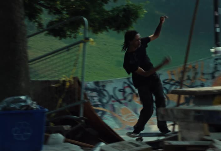 A long-haired man skateboards in slow motion through a gritty, graffiti-covered skate park