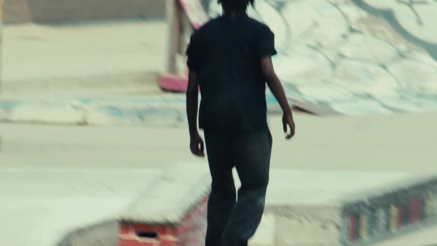 A skateboarder with dreadlocks runs and jumps on their board to ride across the top of a ramp in a skatepark