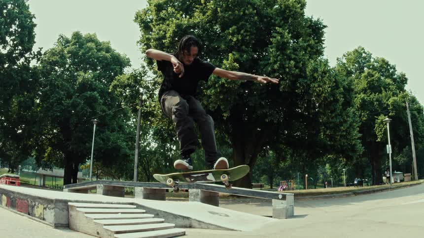 A skateboarder gracefully performs a grind trick down a rail over stairs in a park, captured in stunning slow motion