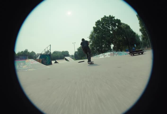 A slow-motion, fisheye lens view follows a skateboarder as they ride through a sunny skatepark and perform a trick