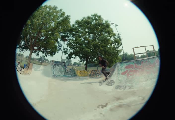 A skateboarder cruises through a concrete skatepark in slow motion and lands a trick on a ramp, captured with a classic fisheye lens