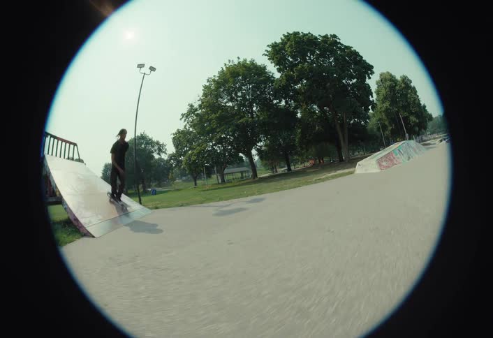 A fisheye, slow-motion shot follows a stylish skateboarder performing tricks throughout an urban skatepark on a sunny day