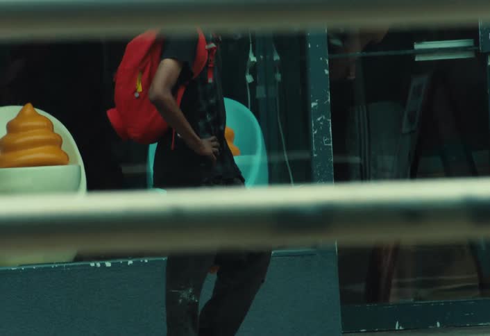A young man with a red backpack and skateboard waits on a city sidewalk as traffic blurs past in the foreground
