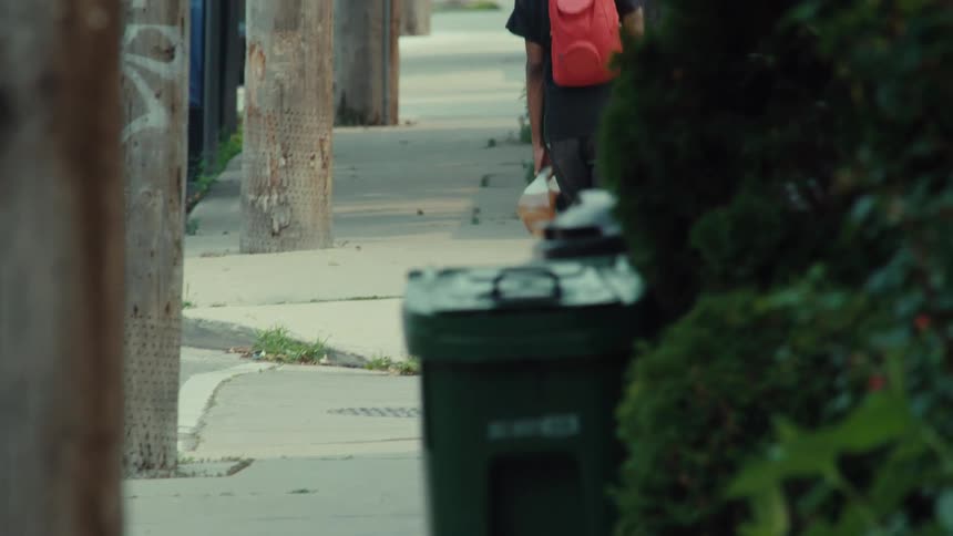 A person with a red backpack skateboards away from the camera down a city sidewalk in slow motion while carrying a jug