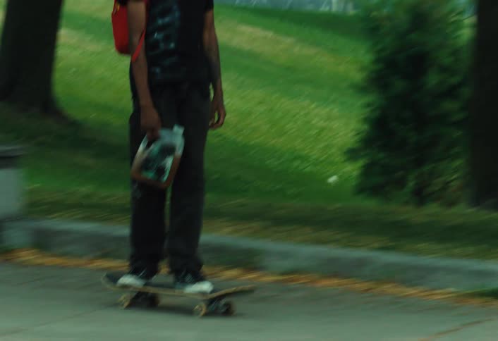 A young man with a red backpack and dreadlocks skateboards down a sidewalk next to a park while carrying a jug