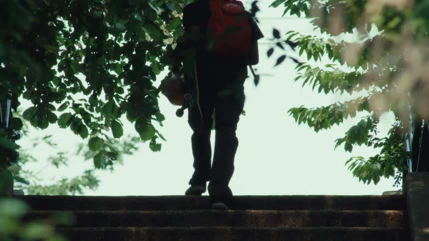 A person carrying a skateboard and a red backpack slowly walks up a long flight of concrete stairs surrounded by lush green trees
