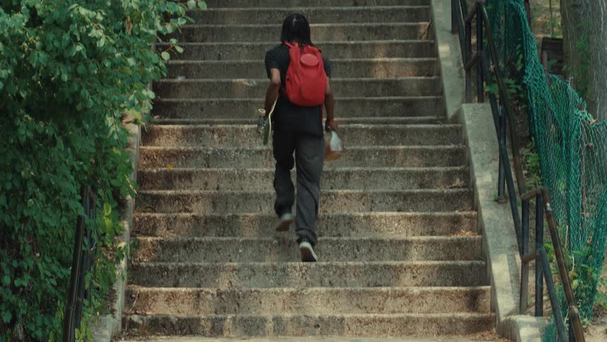 A young Black man with a red backpack carries his skateboard and a jug of liquid as he walks up a long flight of concrete stairs