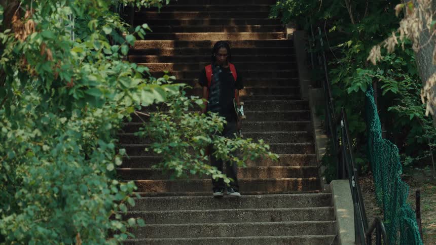 A young Black man with a backpack, skateboard, and glass jug walks down a long flight of outdoor concrete stairs surrounded by lush green trees